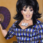 A drag queen holding a fan and wearing a necklace with the letters MKD in the documentary Workhorse Queen