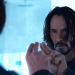 Keanu Reeves looks confused as he holds his fingers up to a mirror in The Matrix Resurrections