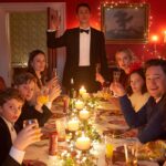 A group of friends and their children sit around a Christmas dinner table, raising their glasses to toast to each other in the movie Silent Night.