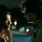 Jared Gilman and Mikey Madison stand in a dark parking lot at night looking into each other's eyes in the movie It Takes Three