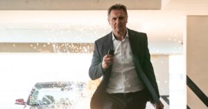 Liam Neeson walks away from an explosion whild holding his phone in his hand in the movie Memory