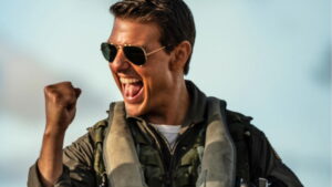 Tom Cruise holds up his fist in triumph and yells while wearing a flight suit and aviators in the movie Top Gun: Maverick