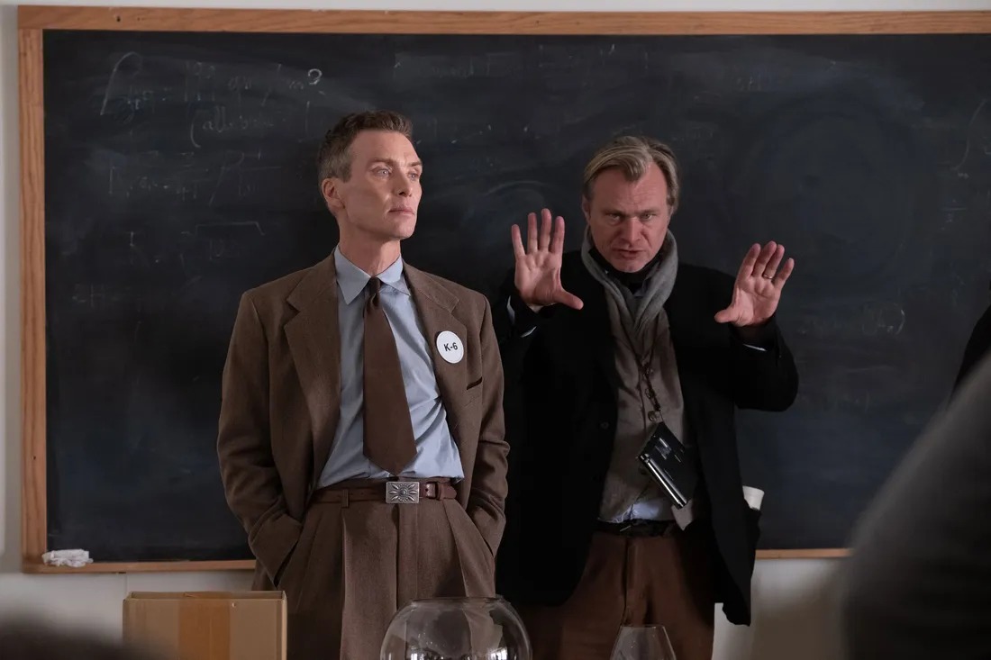 Christopher Nolan stands next to Cillian Murphy in front of a blackboard with his hands out, directing the movie Oppenheimer.