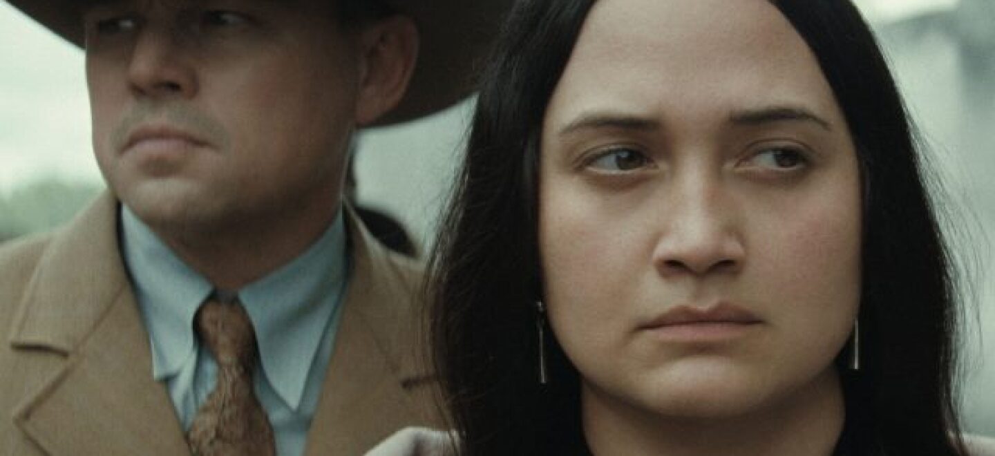 Lily Gladstone looks stoically with a man standing behind her in the movie Killers of the Flower Moon.