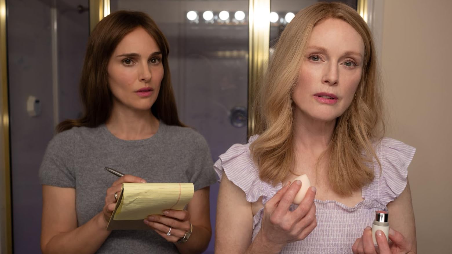 Natalie Portman takes notes while Julianne Moore does her makeup in front of a mirror in the movie May December.