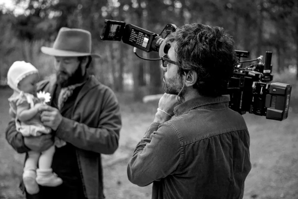 A black and white photo of Adolpho Veloso with a camera looking at Joel Edgerton holding a baby in the film Train Dreams.