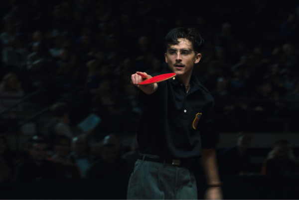 Timothee Chalamet holds a ping pong paddle pointing in the film Marty Supreme.